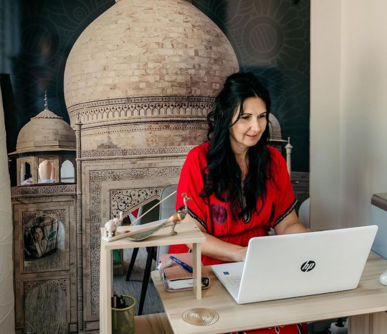 Maitri Ayurvéda központ tulajdonosa dolgozik egy laptopon a héttérben indiai stílusú faldekorációval.